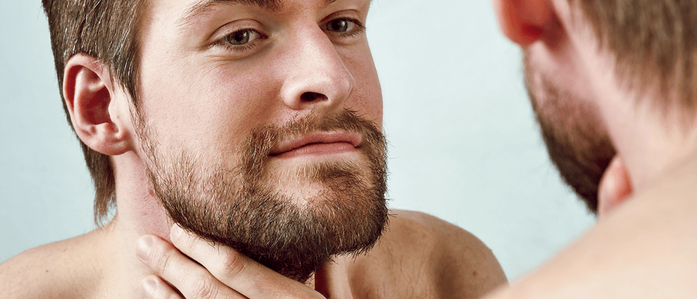 Crescer barba: homem branco mexe e observa sua barba em frente ao espelho.