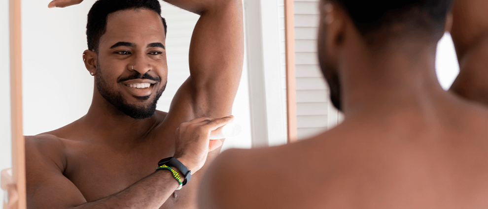 Depilação da axila: homem negro jovem sorridente utliza o auxílio de um aparelho de barbear para remover pelos de sua axila em frente ao espelho.