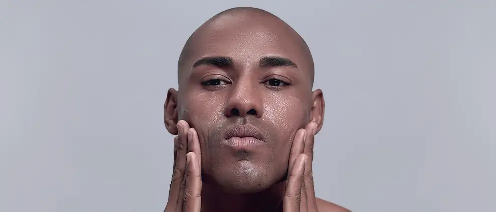 Esfoliante corporal: jovem homem negro com expressão séria usa as duas mãos para espalhar creme pelo rosto.