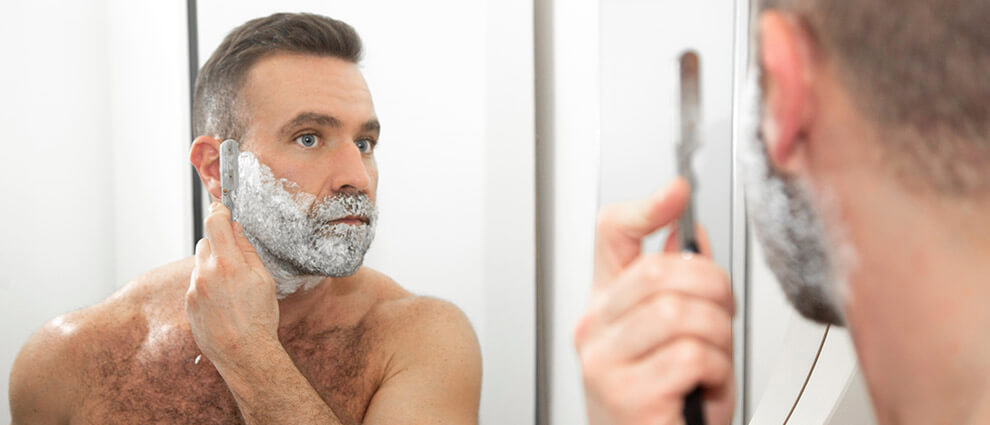 Homem branco, de olhos azuis, topete e sem camiseta, passando a navalha sobre seu rosto coberto de espuma de barbear.
